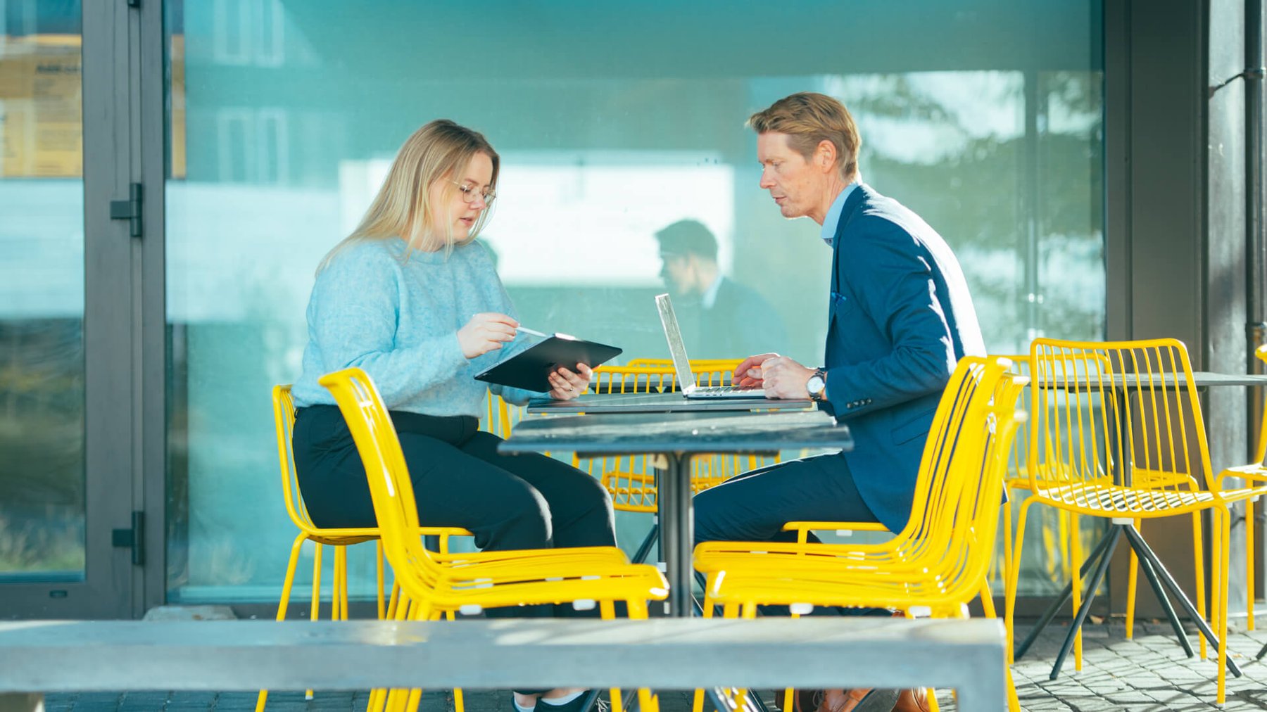 En dame og en mann sitter på en kafe med gule stoler og jobber